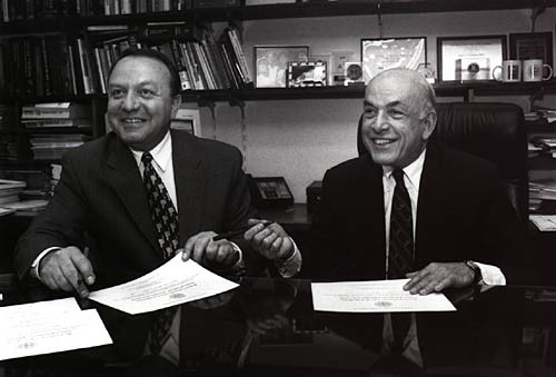 black and white photo of two men seated, wearing suits and ties smiling broadly