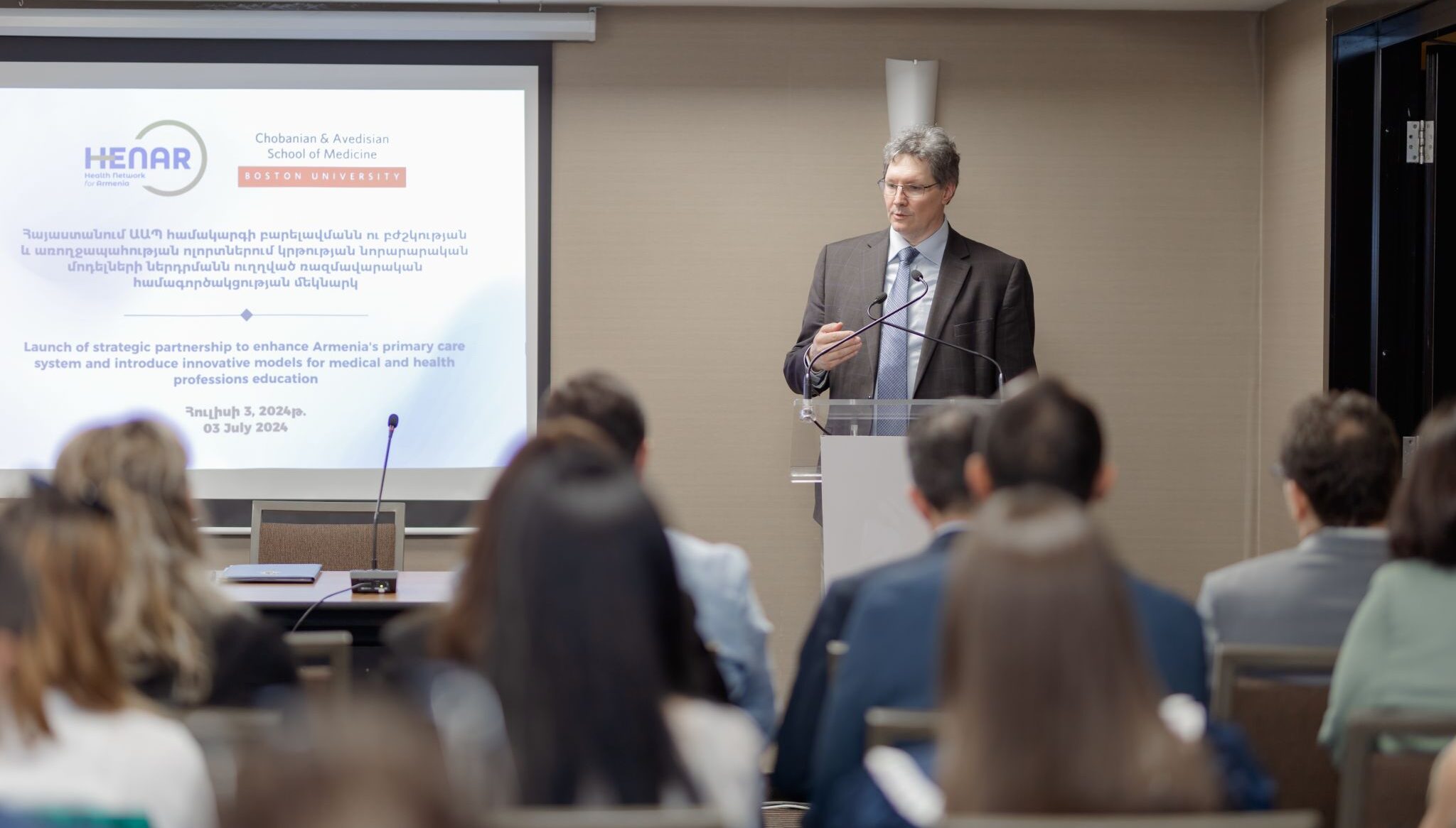 Man in suitjacket standing at podium at front of room beside powerpoint slides