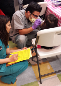 David Garazi DMD 15 gives an oral health screening to a child as Rifat Malhotra AS 15 records the results