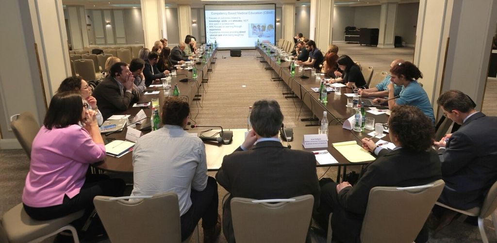 Many people gathered around a large conference room table with powerpoint slides in the distance