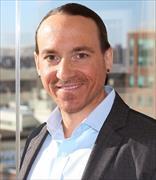 Man with very short dark hair, light facial hair, wearing open neck white button down shirt and dark gray suit jacket, smiling