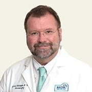 Head and shoulders of man with wireframe eyeglasses with brown hair and facial hair wearing white coat, white shirt and pastel green/blue necktie.