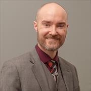 Man with red/brown hair and beard wearing tan suit jacket and vest and burgandy shirt smiling