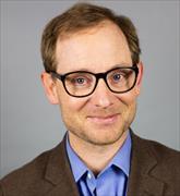 Head and shoulders of man with brown hair, dark rimmed eyeglasses blue shirt and brown suitjacket