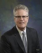 Head and shoulders shot of man with sliver hair, black framed eyeglasses, dark suit jacket, white tie, silver and dark striped tie slightly smiling.