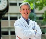 Thomas Perls wearing whitecoat standing outside near large clock, green leaves and building in background