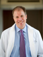 Avrum Spira standing infront of open doors in a hallway wearing a whitecoat, blue shirt, blue/red necktie, smiling broadly