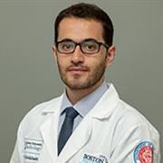 Man with short dark hair, dark facial hair, brown/black eyeglasses, wearing whitecoat