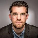 head and shoulders shot of light skin man dark hair on head and facial hair with clear frame eyeglasses, pensive expression