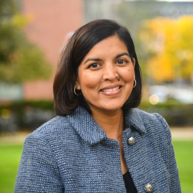 Woman with straight dark brown chin length hair wearing floral top and blue jacket outside in front of green bushes