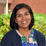 Woman with straight dark brown chin length hair wearing floral top and blue jacket outside in front of green bushes