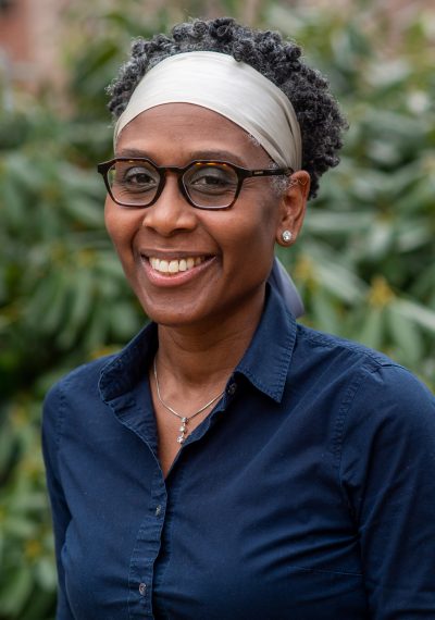 Dr. Coleman in dark blue shirt, light gray headscarf, dark framed eyeglasses, smiling at camera., against a background of green shrubs.