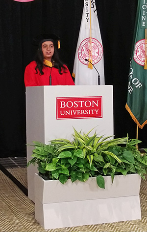 Reshma Gheevarghese in red robe behind white podium giving her address.