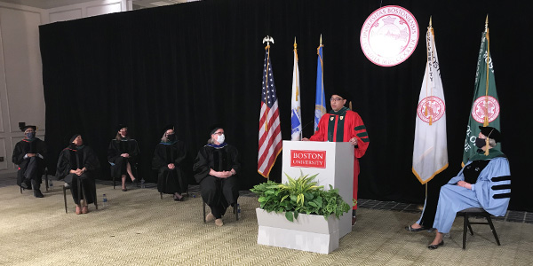 Carlo Pasco in red robe at white podium giving speech surounded by masked faculty seated in a physically distanced manner