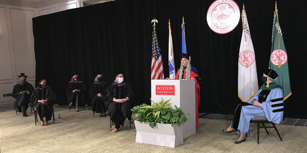 Samantha Shelton in red robe behind white podium giving her speech surrounded by masked faculty seated physically distanced