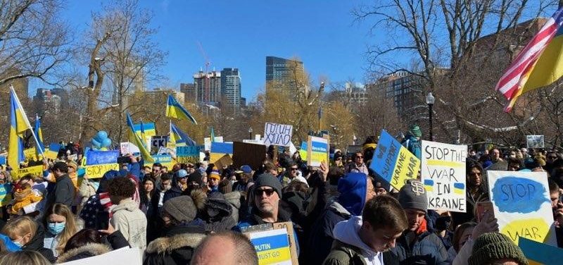 Peace march protesters holding signs, Ukrainian flag, blue balloons, on a winter day with sun shining