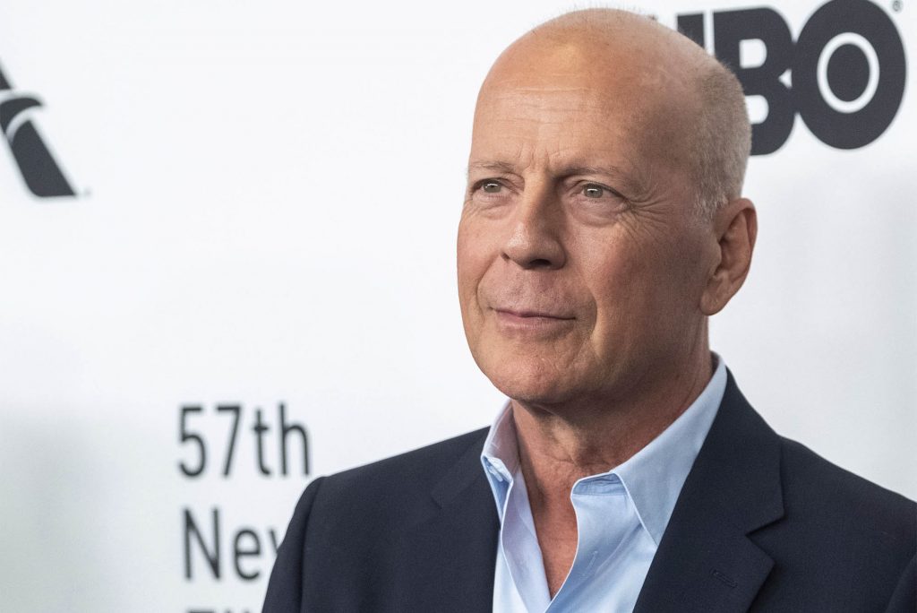 Bruce Willis in a light blue shirt and navy jacketagainst a white background