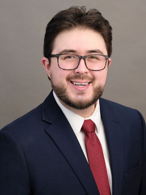 Head and shoulders man with glasses dark hair and beard wearing navy suit, white shirt, red tie