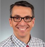 Head and shoulders shot of Claudio Morera wearing a plaid shirt, red tie,smiling broadly wearing black rimmed eyeglasses