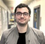 Head and shoulders of Alex Serafini we in black tshirt and gray windbreaker jacket, wearing black framed eyeglasses, dark brown hair, smiling slightly