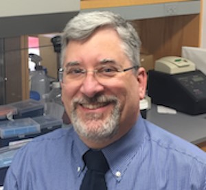 Head and shoulders of Andrew Taylor, blue shirt, dark ties, gray hair and facial hair, eyeglasses smiling 