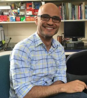 Bald man with dark eyeglasses and plaid shirt smiling, seated with bookcases behind him