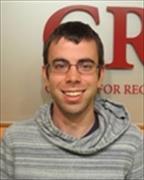 Head and shoulders image of man with short brown hair wearing eye glasses smiling broadly in front of CReM sign on wall.