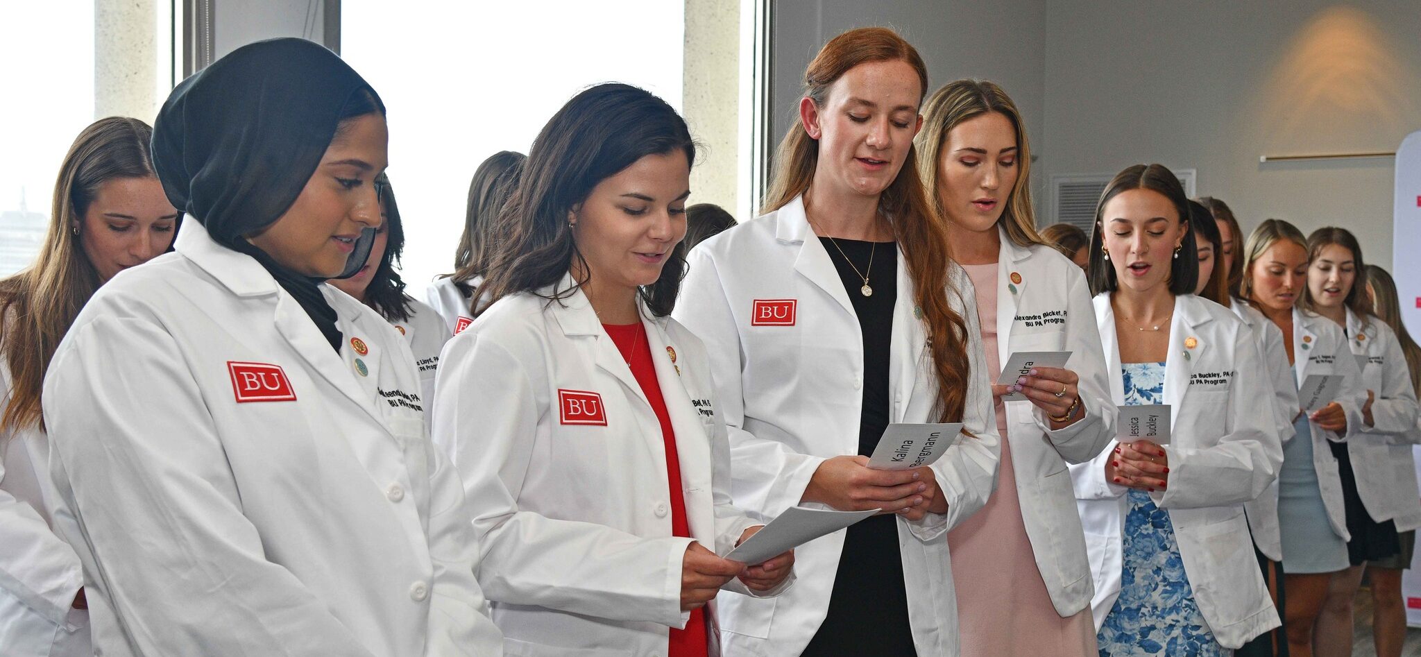 Students standing wearing white coats holding a program from which they are reading the oath