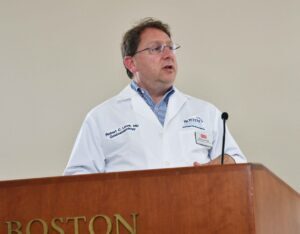 Brownish haired man wearing eyeglasses, white coat speaking behing a wood podium