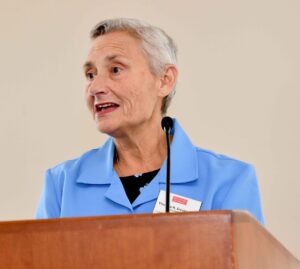 white haired woman blue jacket speaking behind a podium