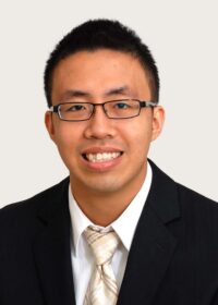 Man wearing eyeglasses, short dark hair, dark suit jacket, white shirt beige necktie smiling broadly