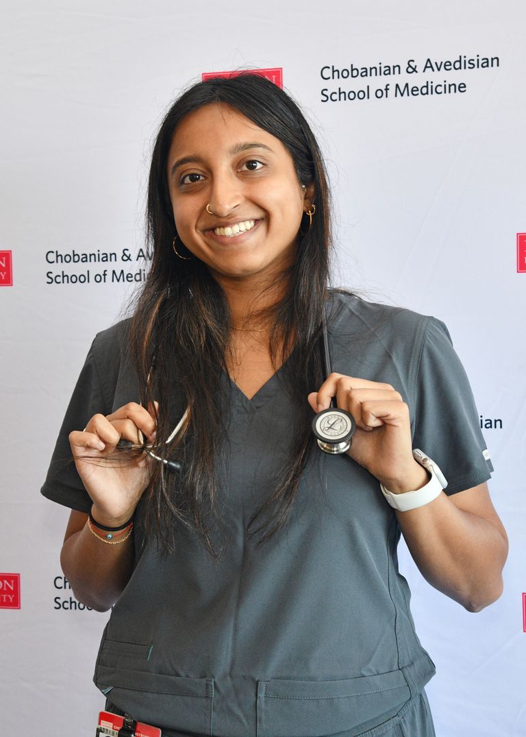 Woman smiling broadly with long brown hair parted in the middle wearing stethoscope wrapped over her shoulders, wearing drak green short sleeved shirt in front of a white panel with black and white text of Chobanian & Avedisian School of Medicine