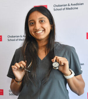 Woman smiling broadly with long brown hair parted in the middle wearing stethoscope wrapped over her shoulders, wearing drak green short sleeved shirt in front of a white panel with black and white text of Chobanian & Avedisian School of Medicine