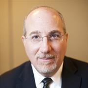 Head and shoulders of man with gray beard and mustache wearing wire frame eye glasses, dark suit jacket, white shirt, dark ties smiling gentl