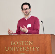 Man in pink v-neck sweater with dark eyeglasses, hair and beard standing behind podium presenting