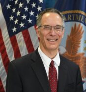 Man with gray hair and glasses wearing blue suit, white shirt, red tie, infront of US flag and veteran flag