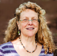 Woman with long blond wavy/curly hair, frameless eyeglasses, wearing striped purple top and multicolored pearl necklace, smiling