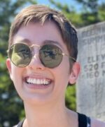 Woman with short brown hair, gold wire frame round sunglasses with green lenses smiling broadly