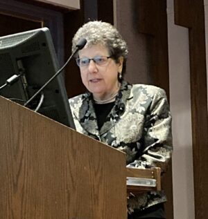 Woman standing behind podium with microphone and computer monitor visible