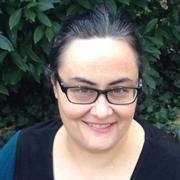 head and shoulders of woman with dark hair and dark rimmed eyeglasses standing in front of a green bush outside