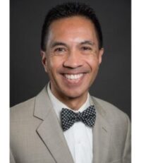 man with dark short hair smiling broadly wearing white shirt, dark bow tie, taupe suit jacket