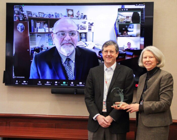 Man with white beard and eyeglasses on large video screen behind Drs. Jack and Antman in foreground holding a crystal award honoring Project RED
