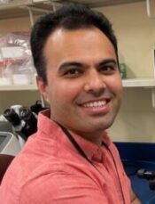 Man with short dark hair smiling, wearing coral colored shirt with microscope behind him.