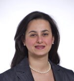 head and shoulders of woman with long dark hair wearing dark suit jacket and pearl necklance and earrings, smiling