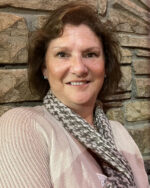 Woman in front of stone wall, brown wavy hair, tan shirt, patterned scarf around neck.