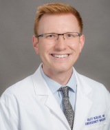 Riley Kolus in white coat, light blue shirt, gray neck tie, wearing eyeglasses smiling broadly