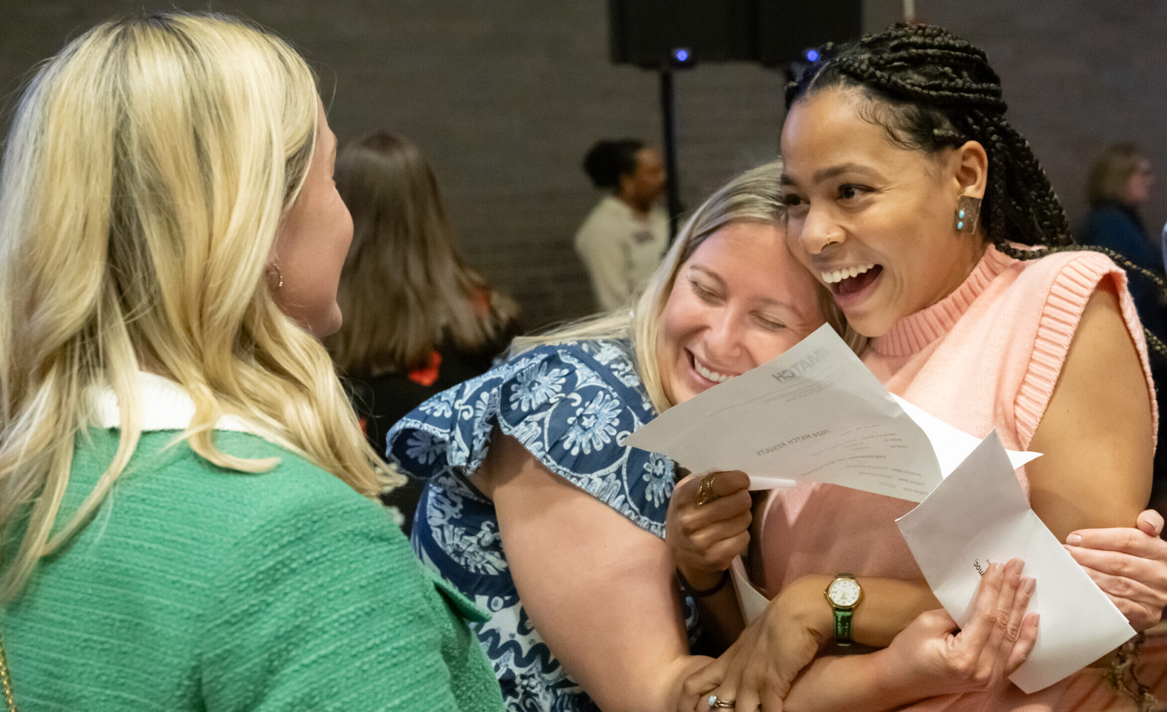 three woman, two embracing while smiling holding match letter