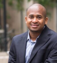 Dr. Jones standing outside in a medium blue suit jacket, light blue open neck dress shirt, smiling,