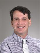 head and shoulders of man with dark hari wearing checked shirt light tie, smiling