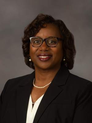 Head and shoulders picture of Loretta Jackson-Williams wearing dark suit jacket, white blouse, strand of pearls. Chin length dark hair, eyeglasses, smiling